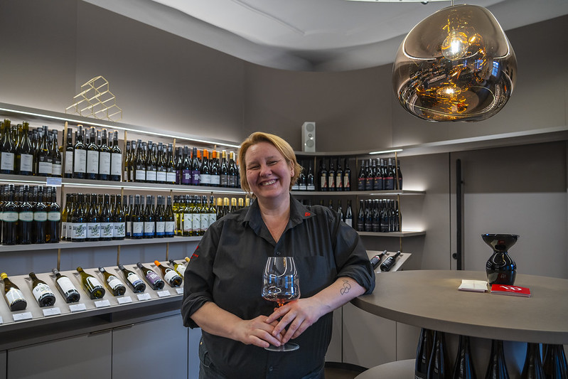 Lachende blonde vrouw met een glas wijn in haar hand en op de achtergrond kasten met wijnflessen.