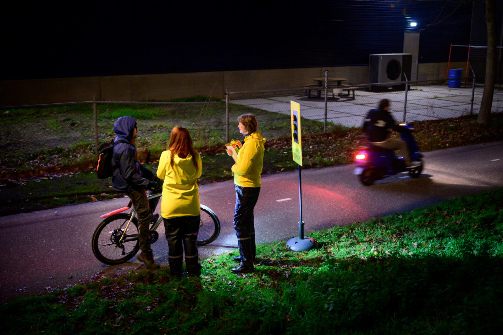 Stoplicht voor mannen 15 Twee vrouwen met gele jassen praten met een mountainbiker op een donker fietspad.