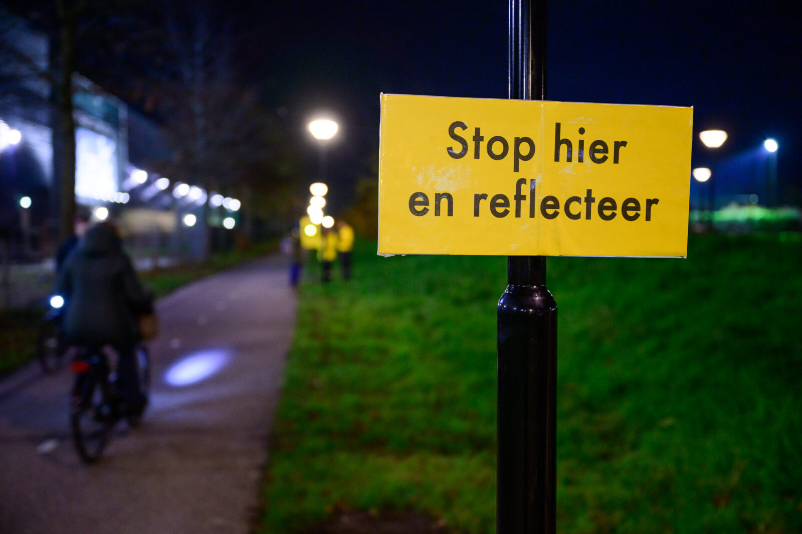 Stoplicht voor mannen 14 Bordje 'Stop hier en reflecteer' op een paal langs een donker fietspad.